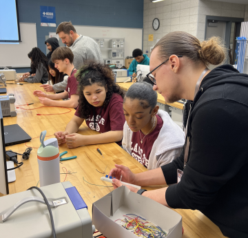 Several students sit at a workbench assembling electronics while an instructor offers guidance in a classroom lab setting.