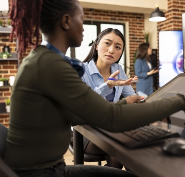 Two colleagues collaborate at a workstation in a modern office, discussing a 3D environment displayed on a large angled touchscreen monitor while another teammate works in the background.