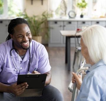 A smiling healthcare worker in purple scrubs talks with an elderly woman using a walker