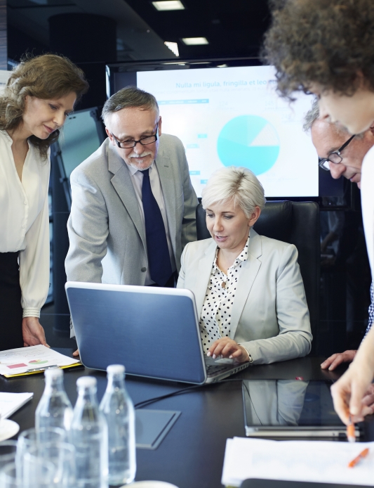 Five professionals gather around a laptop in a conference room, reviewing information together